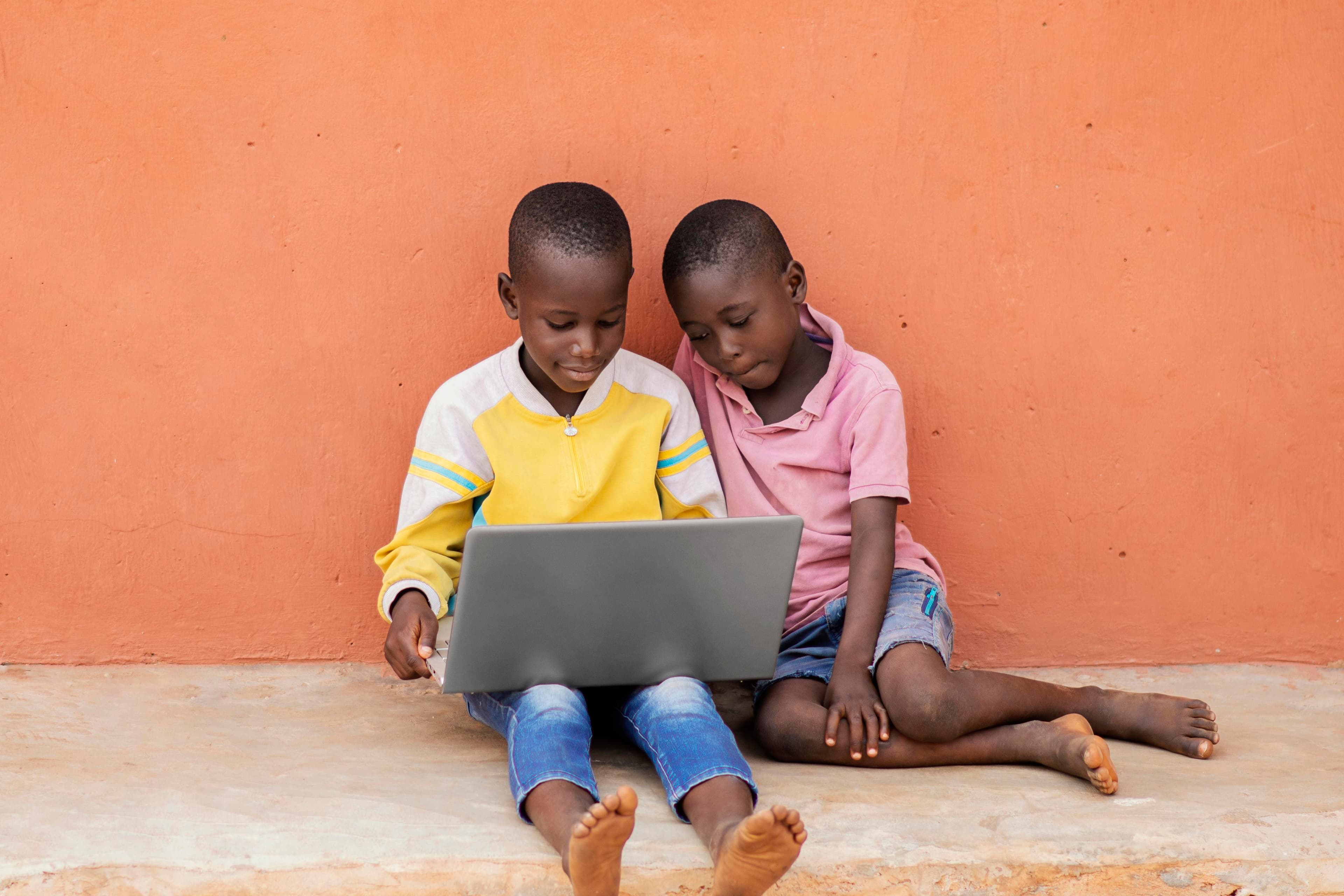 Two young students sharing a laptop, sitting against an orange wall.